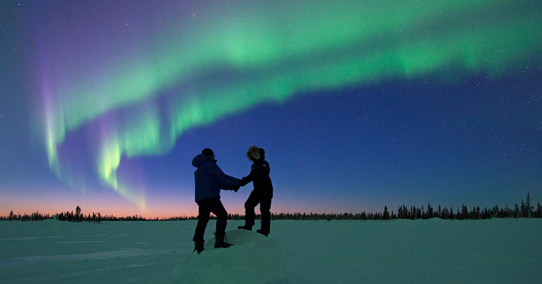 two people holding hands on the snow with the northern lights showing above